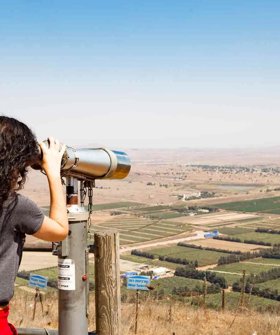 מטיילת צופה בנוף רמת הגולן והכנרת דרך משקפת בנקודת תצפית.